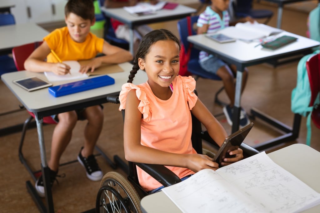 young girl in wheelchair sits at desk in classroom. ableism concept