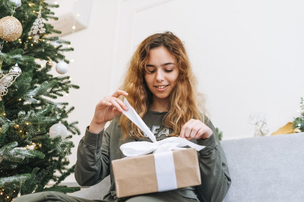 Teenage Girl With Curly Hair Holds Gift Box In Her Arms While Sitting On The Sofa In The Living Room With A Christmas Tree. Happy Teenager In The Decorated Living Room At House On Christmas Morning