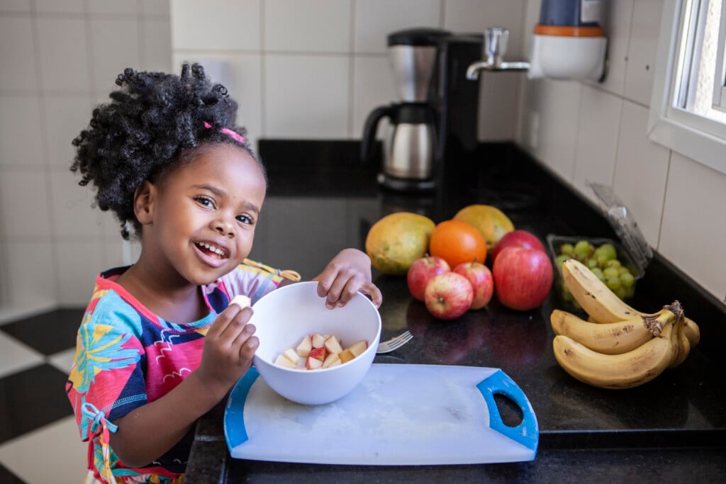 A toddler holds a bowl of chopped apples on a kitchen counter and smiles at the camera
