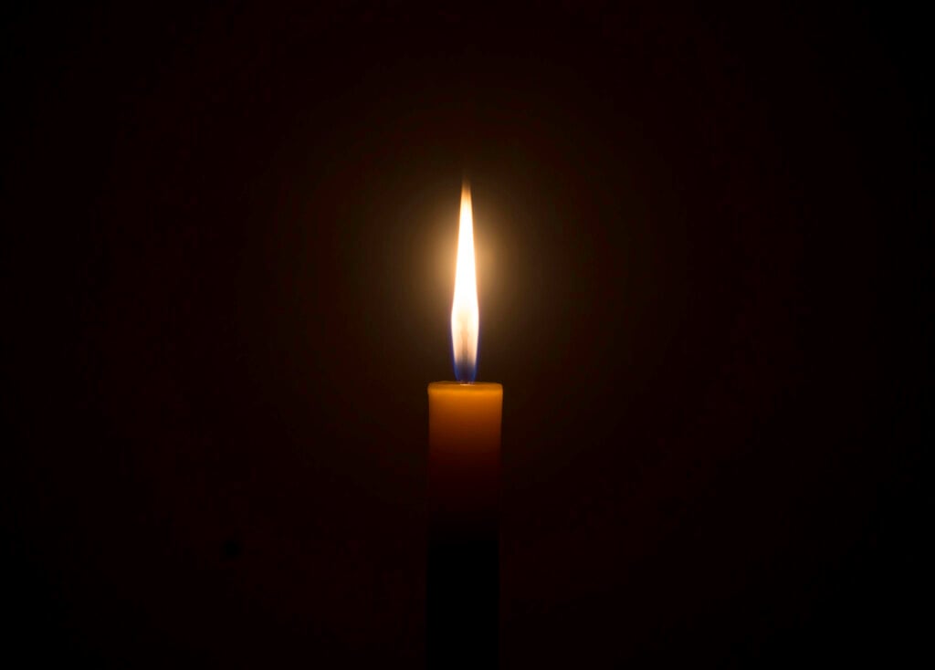 A Single Burning Candle Flame Or Light Glowing On An Orange Candle On Black Or Dark Background On Table In Church For Christmas, Funeral Or Memorial Service With Copy Space