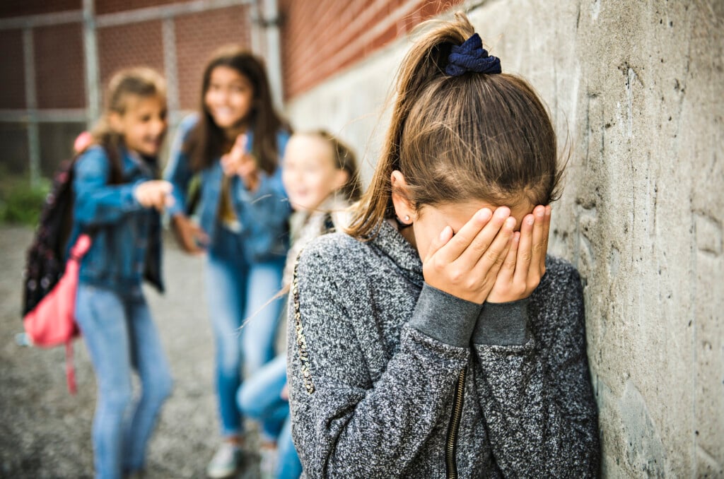 a young student with their head in their hands, three other students laughing at them in the background, bullying concept