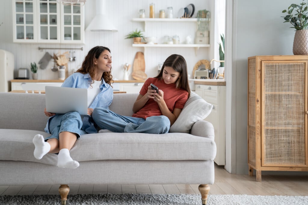 a mom on a laptop tries to look at her teen daughter's phone, as the teen leans away