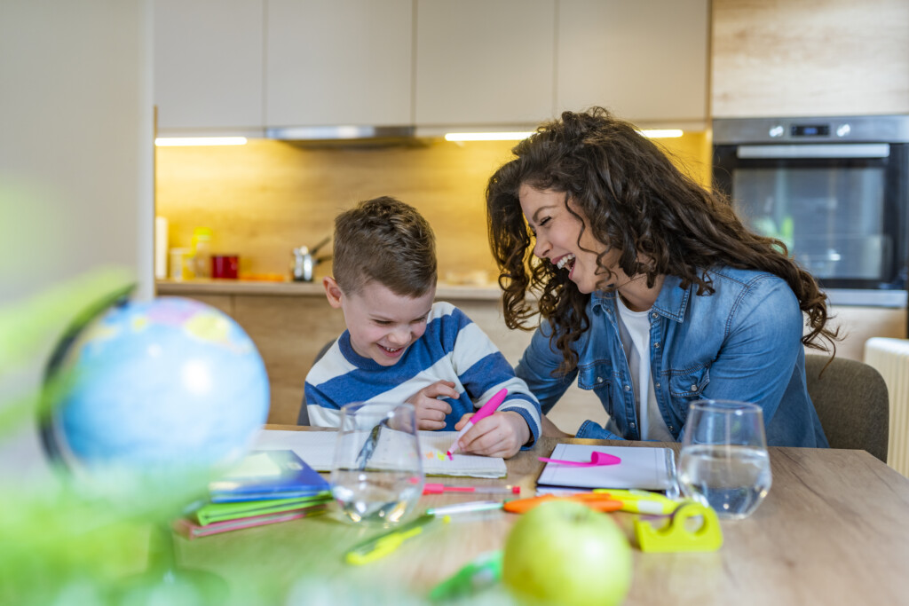 Young Mother Sitting At A Table At Home Helping Her Young Son With His Homework