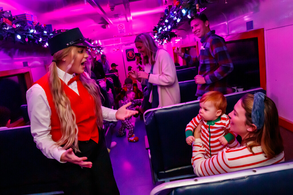 train conductor dressed in red vest talks to mother and baby in red and white striped pajamas