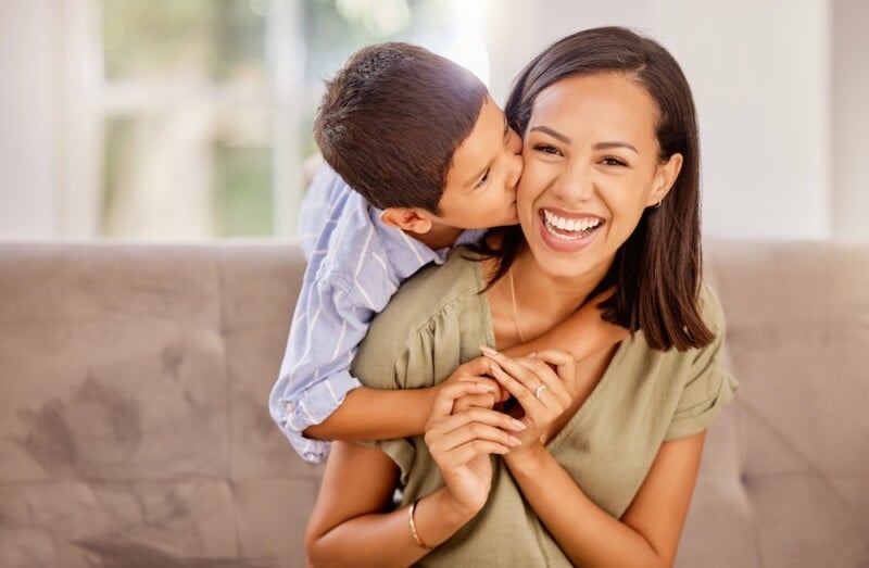 young child hugs and kisses mom from behind as they sit on a coach