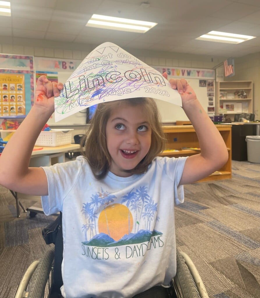 a young child in a wheelchair in a classroom, smiling with a paper hat