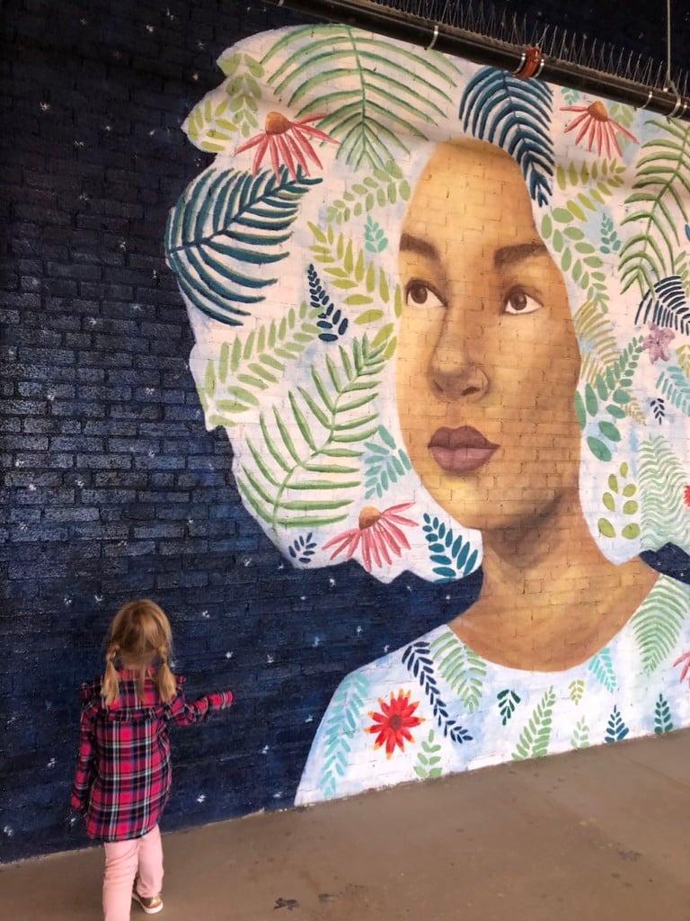 a young girl stands in front of a mural at mother road market