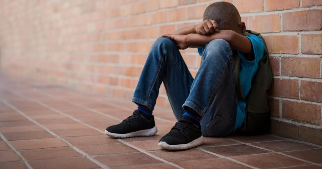 Young boy sitting alone with sad feeling at school. Depressed african child abandoned in a corridor and leaning against brick wall. Bullying