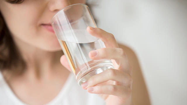 Happy beautiful young woman drinking water. Smiling caucasian female model holding transparent glass in her hand. Closeup. Focus on the arm