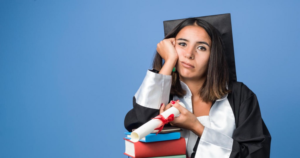 Young beautiful mixed race woman wearing a black graduation gown and cap looks worried while holding her diploma in left hand.She is holding her hand in her hand.She contemplating about her future.The background is blue.Shot in studio with medium format camera Hasselblad.