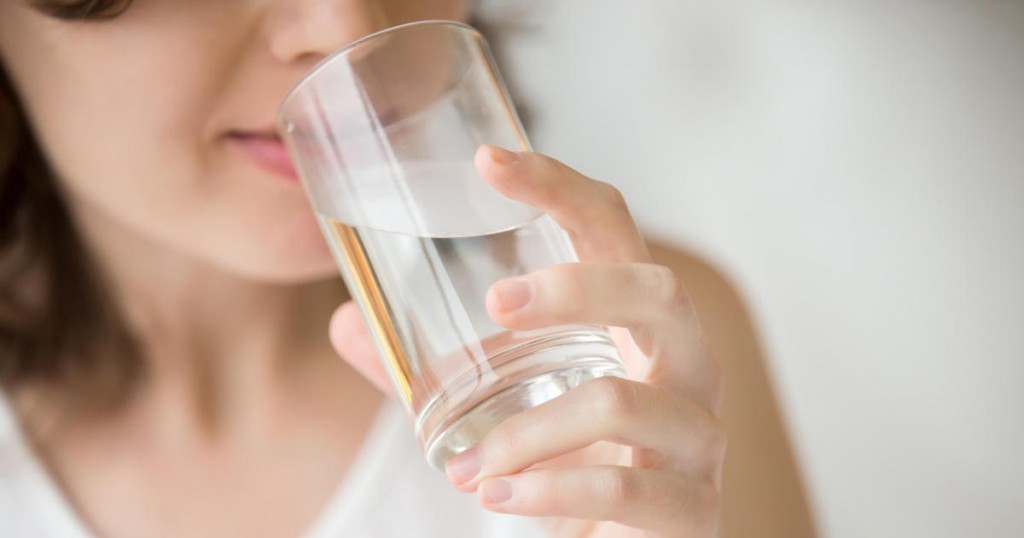Happy beautiful young woman drinking water. Smiling caucasian female model holding transparent glass in her hand. Closeup. Focus on the arm