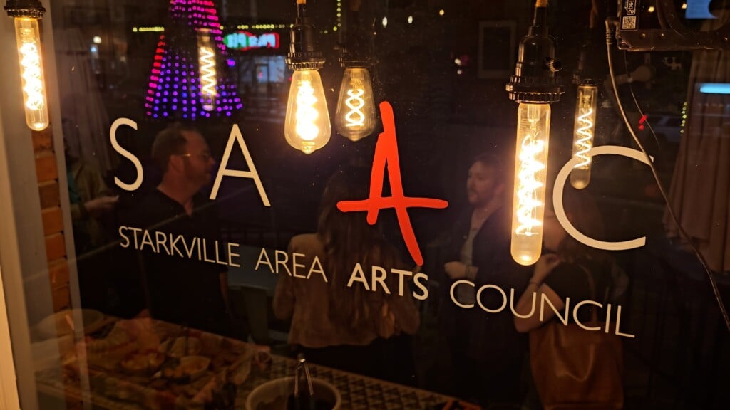 Starkville Area Arts Council Logo On Glass Behind Edison Bulbs With People On The Other Side Of The Glass