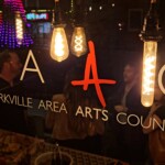 Starkville Area Arts Council Logo On Glass Behind Edison Bulbs With People On The Other Side Of The Glass