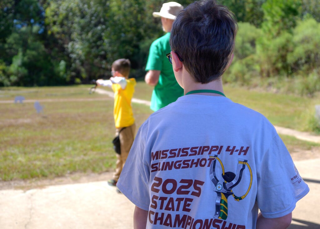 In the foreground, a boy pictured from behind wearing a T-shirt reading “Mississippi 4-H Slingshot 2025 State Championship” looks on as another boy aims at a target with a slingshot.