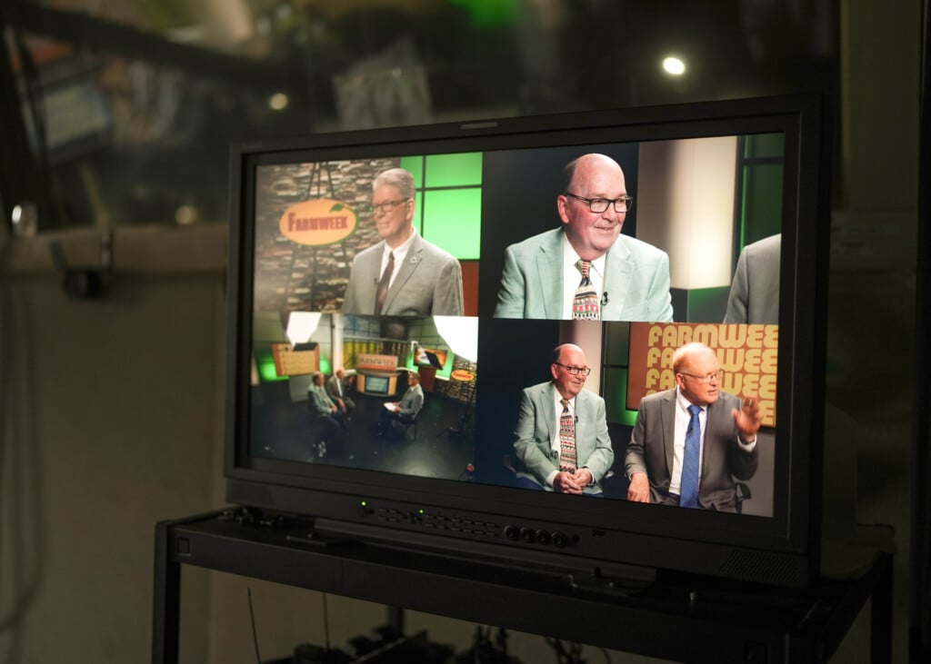 Video monitor displays a talk show set with two men in suits being interviewed, shown from multiple camera angles, with a "Farmweek" sign in the studio background.