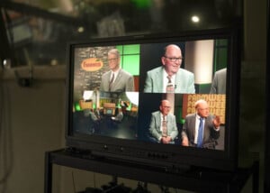 Video monitor displays a talk show set with two men in suits being interviewed, shown from multiple camera angles, with a "Farmweek" sign in the studio background.