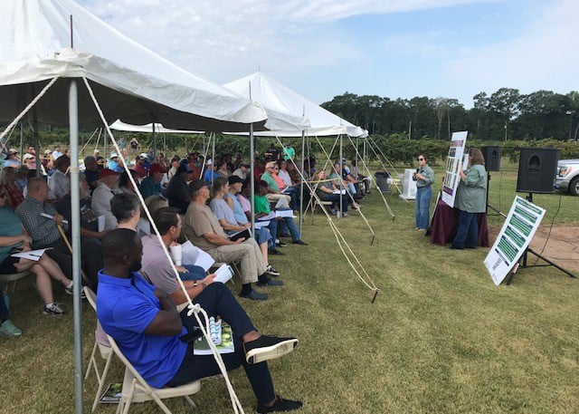 Presenters address a large crowd sitting under outdoor tents.