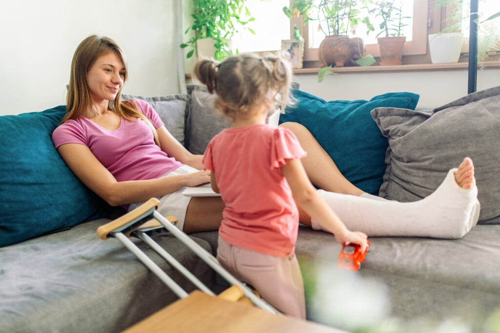 Mother With Cast On Her Leg Working On Her Laptop And Her Young Daughter Playing