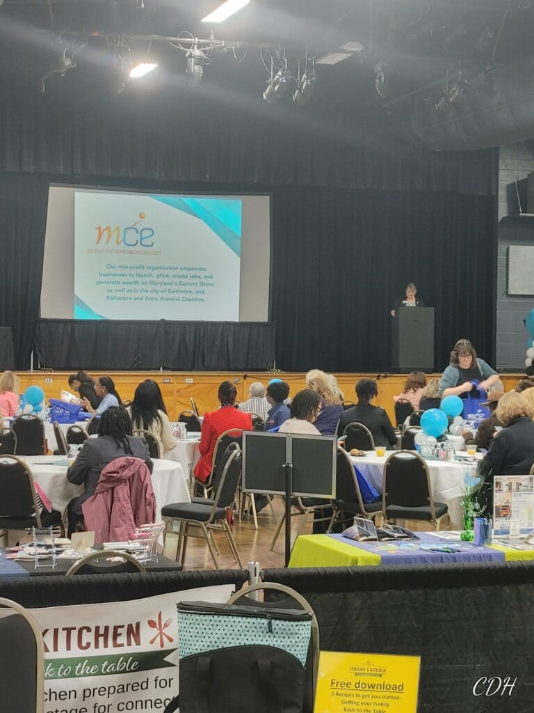 Women attendees sit in an auditorium as a presenter is on stage by a podium presenting during a Maryland Capital Enterprises Women's Business Center conference.