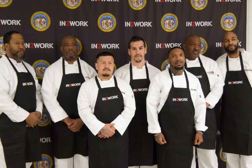 Inmates at Eastern Correctional Institution stand in front of the IN2WORK and Maryland Department of Public Safety and Correctional Services backdrop.