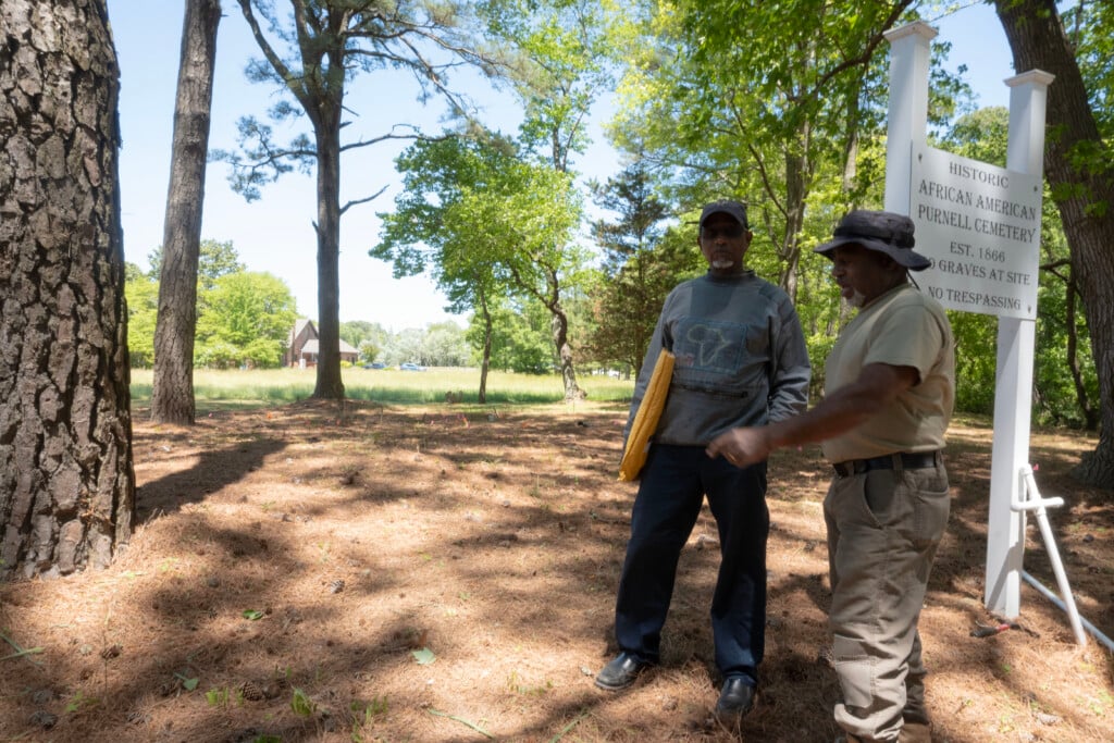 Bobby Purnell and Al Showell at the Purnell family cemetery, standing in front of a sign in front of a field with little colored flags, marking locations of graves.