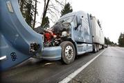 A semi-truck with visible damage to its hood sits parked on the shoulder just ahead of a car covered in mud and debris and with a smashed windshield just beyond where an Amtrak train lay spilled onto Interstate 5 below as some train cars remain on the tracks above Monday