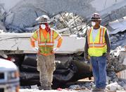 Workers stand in front of crushed cars under a section of a collapsed pedestrian bridge