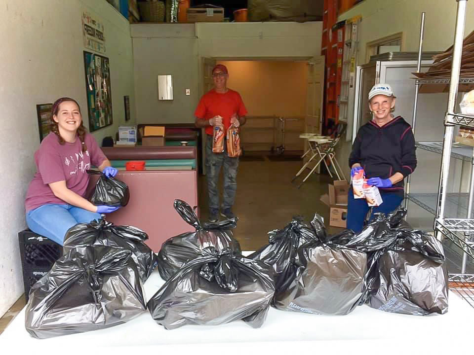 One woman and two men stand behind a group of garbage bags filled with food.